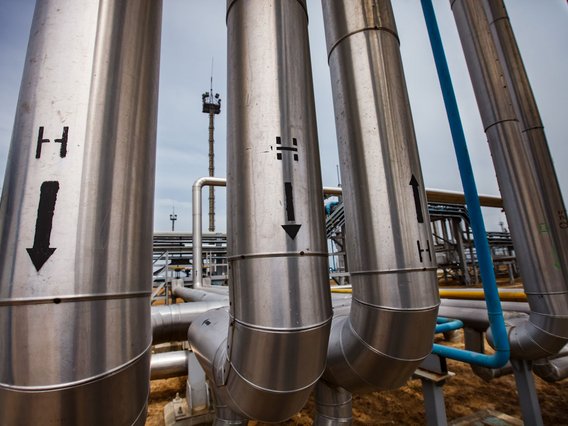 Wasserstoffanlage große, silberne Rohrleitungen in einer Industrieanlage. Auf den Rohren sind schwarze Pfeile und das Symbol „H“ zu sehen, die vermutlich den Fluss von Wasserstoff kennzeichnen.