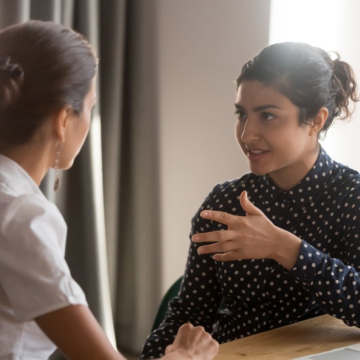 Gesprächssituation Mentoring: Zwei Frauen sitzen einander zugewandt an einem Tisch. Die eine Frau erklärt der anderen etwas.