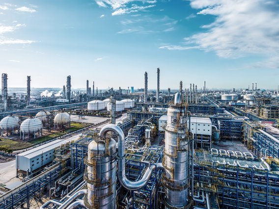 Große Industrieanlage mit Türmen, Tanks und verbundenen Rohrleitungen unter einem strahlend blauen Himmel, mit Grünflächen im Hintergrund.
