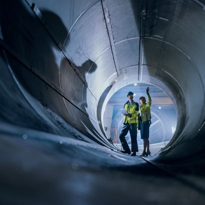 Wärmenetz_Rohr Two Heavy Industry Engineers Walking Inside Pipe, Use Laptop, Have Discussion, Checking Design.