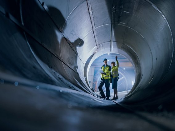 Wärmenetz_Rohr Two Heavy Industry Engineers Walking Inside Pipe, Use Laptop, Have Discussion, Checking Design.