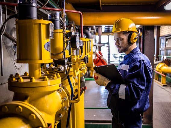Person in Arbeitskleidung, die einen gelben Schutzhelm, Gehörschutz und rote Handschuhe trägt. Die Person steht in einer Industrieanlage und prüft gelbe Maschinen oder Rohrleitungen, während sie ein Klemmbrett in der Hand hält.