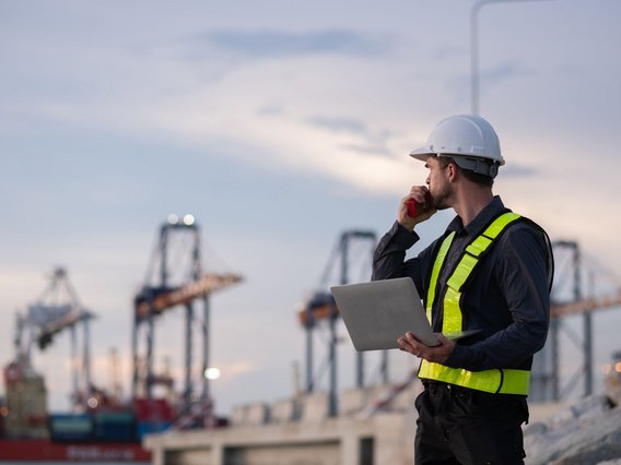 Hafen_Arbeiter_Kräne Person mit Schutzhelm und gelber Warnweste, die auf einem Industriegelände steht. Die Person hält ein Funkgerät und einen Laptop in den Händen und blickt zur Seite. Im Hintergrund sind unscharf große Hafenkräne und Container zu sehen