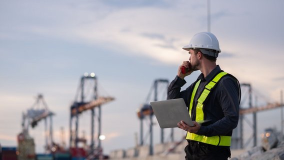 Hafen_Arbeiter_Kräne Person mit Schutzhelm und gelber Warnweste, die auf einem Industriegelände steht. Die Person hält ein Funkgerät und einen Laptop in den Händen und blickt zur Seite. Im Hintergrund sind unscharf große Hafenkräne und Container zu sehen