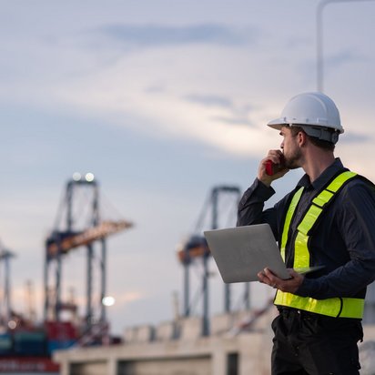 Hafen_Arbeiter_Kräne Person mit Schutzhelm und gelber Warnweste, die auf einem Industriegelände steht. Die Person hält ein Funkgerät und einen Laptop in den Händen und blickt zur Seite. Im Hintergrund sind unscharf große Hafenkräne und Container zu sehen