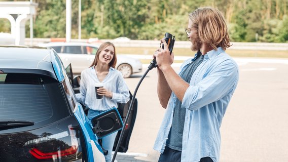 E-Auto Ein Mann steht am E-Auto und hält das Ladekabel. Eine Frau lehnt an der Beifahrertür und schaut zu.