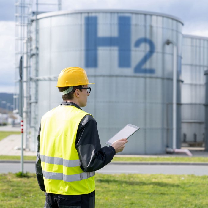 Person mit gelbem Schutzhelm und gelber Warnweste, die auf einem Industriegelände steht. und auf Wasserstofftanks blickt.