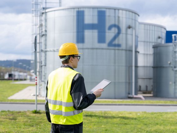 Wasserstofftank Person mit gelbem Schutzhelm und gelber Warnweste, die auf einem Industriegelände steht. und auf Wasserstofftanks blickt.