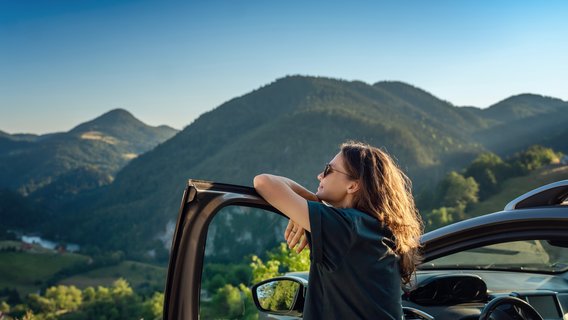 Urlaub Eine Frau steht an der offenen Fahrertür ihres Autos. Sie stützt die Arme auf dem Rahmen der Tür ab und schaut in Richtung Berge.