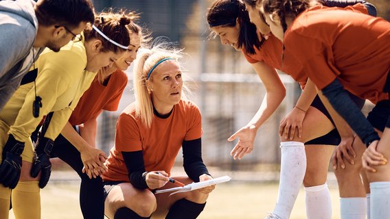 Fußball Fußballerinnen in rot-schwarzen Trikots stehen um ihre Trainerin herum. Sie hat ein Klemmbrett in der Hand.