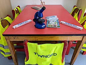 A microscope and other equipment on a red table. Children's high visibility vests with TÜV NORD GROUP logo hang on the chairs.