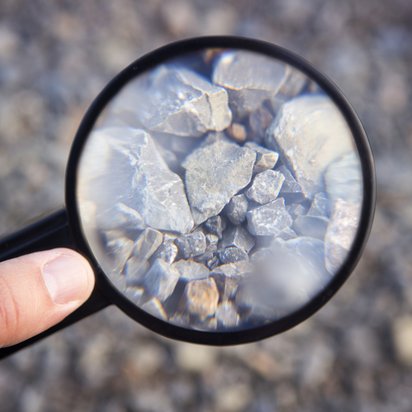 Lagerstättenexploration und Ressourcenabschätzung Symbolbild für Lagerstättenexploration und Ressourcenabschätzung: eine Hand hält eine Lupe über eine Gesteinsprobe und vergrößert einen Teil davon.