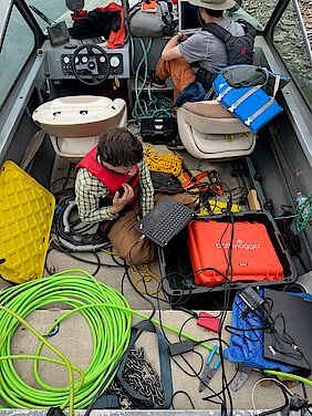 a scientist sitting in a speedboat with a laptop on his knee, surrounded by equipment and cablesaptop