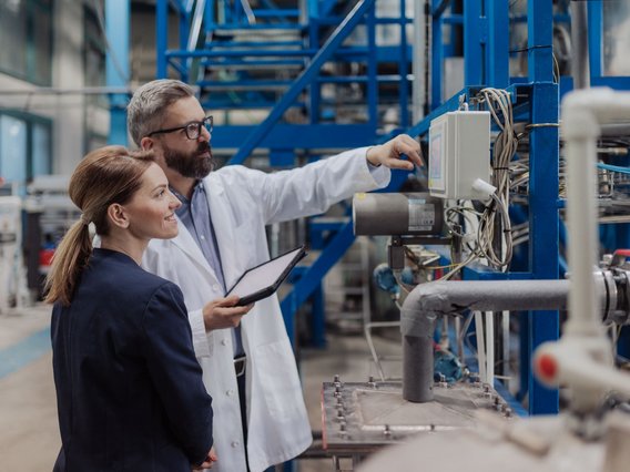 Industrienanlage_Erklärung_Rohre Zwei Personen in einer industriellen Umgebung. Eine Frau im Business-Outfit und ein Mann im weißen Laborkittel stehen vor einer technischen Anlage mit vielen Rohren und Kabeln.