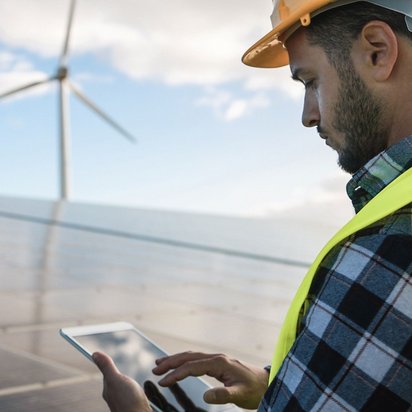 PV_Windenergie_Bauarbeiter Das Bild zeigt einen Mann mit Schutzhelm und Warnweste, der ein Tablet benutzt. Im Hintergrund sind große Solarpaneele und zwei Windkraftanlagen zu sehen.