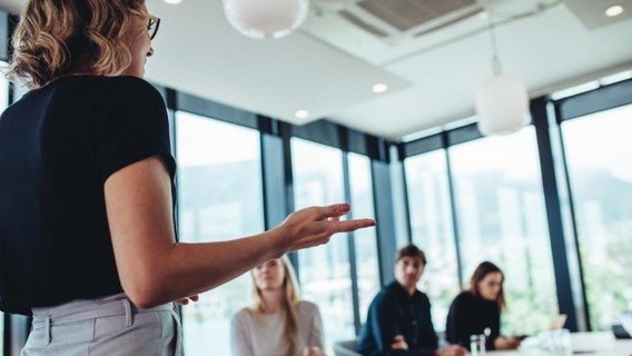 Fortbildung Eine Frau steht vor einem Tisch, an dem weitere Personen sitzen. Sie hält einen Vortrag. Die anderen Personen sind ihr zugewandt und hören zu.