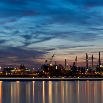 Industrieanlage_Hafen Industrieanlage oder einen Hafen bei Dämmerung. Im Vordergrund spiegelt sich das Licht der Anlage im ruhigen Wasser. Im Hintergrund sind mehrere große Kräne, Schornsteine und beleuchtete Gebäude zu sehen.