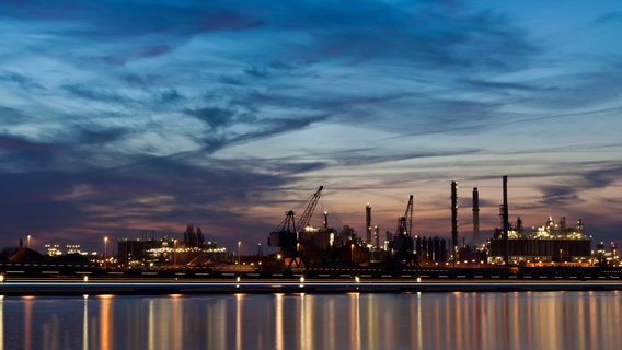Industrieanlage_Hafen Industrieanlage oder einen Hafen bei Dämmerung. Im Vordergrund spiegelt sich das Licht der Anlage im ruhigen Wasser. Im Hintergrund sind mehrere große Kräne, Schornsteine und beleuchtete Gebäude zu sehen.