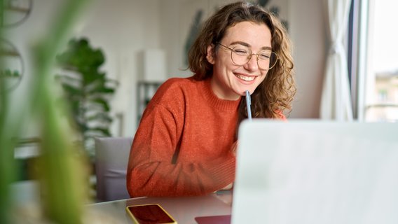 eLearning Eine junge Frau sitzt vor ihrem Laptop am Schreibtisch. Sie tippt sich mit einem Stift ans Kinn.