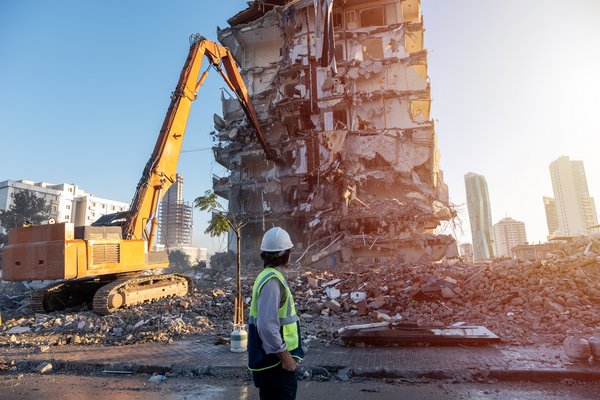 Ingenieur auf einer Baustelle, im Hintergrund reißt ein Bagger ein altes Gebäude ab, von dem nur noch offene Stahlbetonstrukturen zu sehen sind..