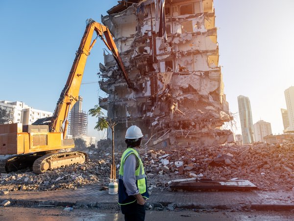 Ingenieur auf einer Baustelle, im Hintergrund reißt ein Bagger ein altes Gebäude ab, von dem nur noch offene Stahlbetonstrukturen zu sehen sind..