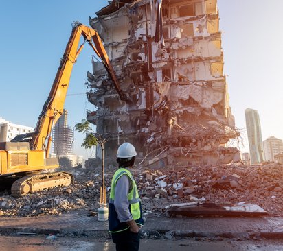 Rückbaukonzepte Ingenieur auf einer Baustelle, im Hintergrund reißt ein Bagger ein altes Gebäude ab, von dem nur noch offene Stahlbetonstrukturen zu sehen sind..