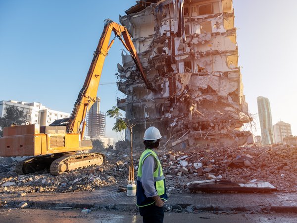 Ingenieur auf einer Baustelle, im Hintergrund reißt ein Bagger ein altes Gebäude ab, von dem nur noch offene Stahlbetonstrukturen zu sehen sind..