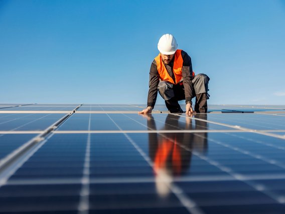 PV-Anlage_Arbeiter Das Bild zeigt eine Person mit Schutzhelm und orangefarbener Warnweste, die auf einem Solarpanel arbeitet.