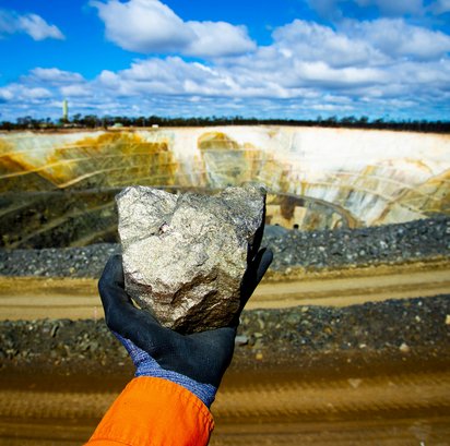 Bergbau & Rohstoffe Symbolbild für den Bereich Bergbau & Rohstoffe: eine Hand hält ein Stück Mineralerz vor einem offenen Tagebau in die Höhe