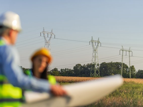 Symbol zum Thema Wegerechtliche Planungsbegleitung: Ingenieure breiten unter freiem Himmel in der Nähe von Stromleitungsmasten einen großen Plan aus