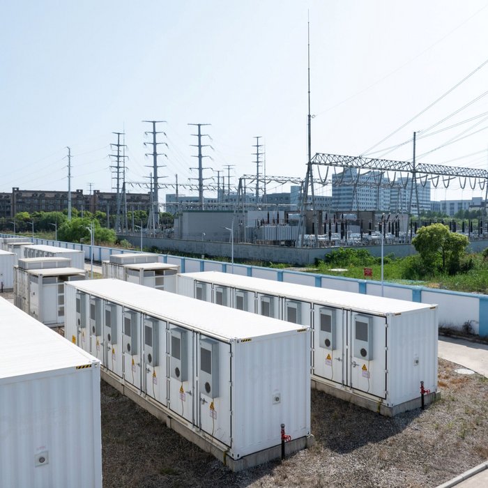 Das Bild zeigt mehrere weiße Container, die als Batteriespeicher dienen und nebeneinander auf einem Gelände stehen. Im Hintergrund sind Stromleitungen, ein Umspannwerk und moderne Gebäude zu sehen. 