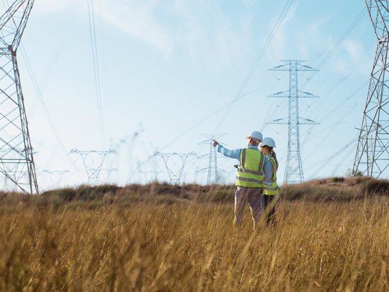 Symbolbild zum Thema Wegerechtliche Baubegleitung: Zwei Ingenieure stehen in einem Feld unterhalb von Stromleitungsmasten