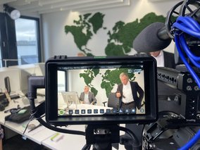 Boris Dombrowski und Knut Hirsch sitzen vor einer Wand mit der Weltkarte. Sie sind durch das Display einer Kamera zu sehen.