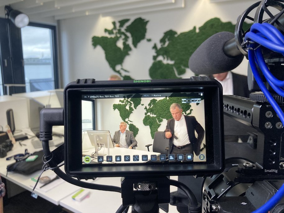 Boris Dombrowski und Knut Hirsch sitzen vor einer Wand mit der Weltkarte. Sie sind durch das Display einer Kamera zu sehen.