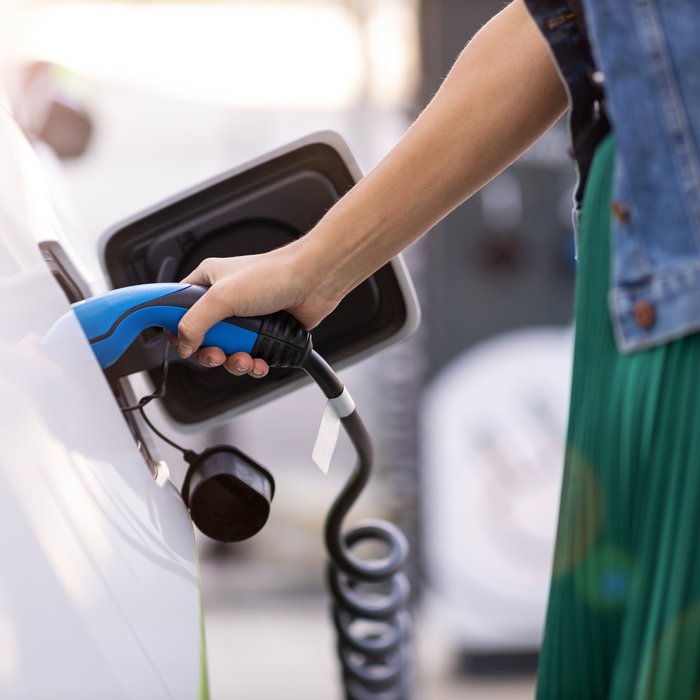 Person, die ein Elektroauto an einer Ladestation auflädt. Die Hand hält einen blauen Ladestecker, der in die Ladebuchse des Fahrzeugs gesteckt wird. 