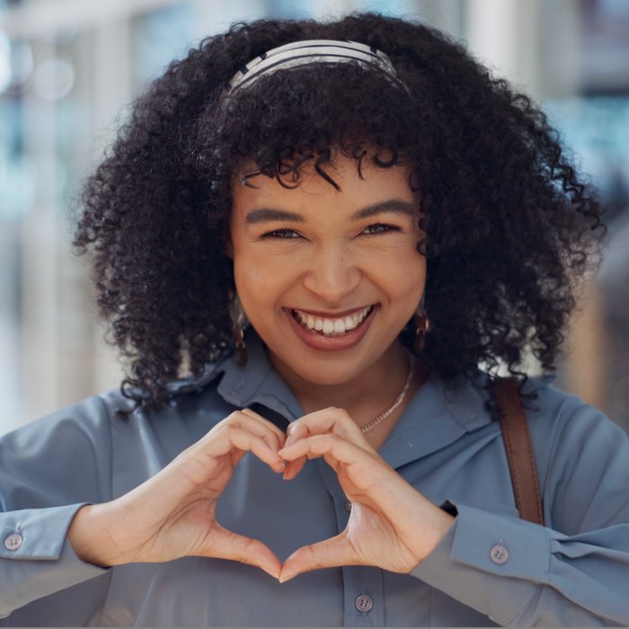 Junge Frau lacht strahlend in die Kamera formt ein Herz mit Ihren Händen