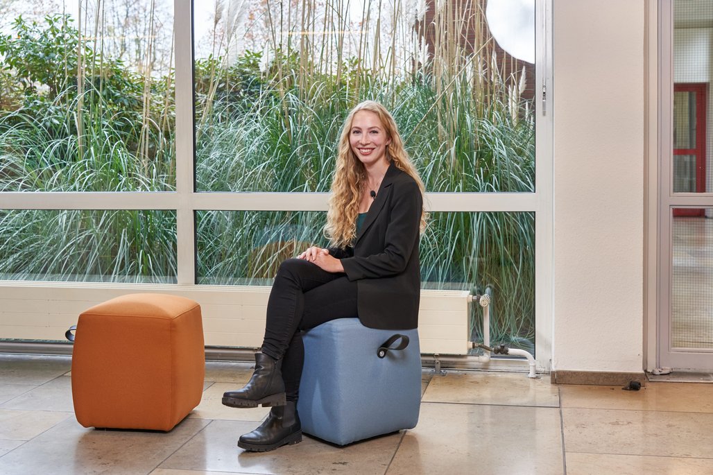 Alisha Gertack sitzt auf einem blauen Sitzhocker vor einer Fensterfront.