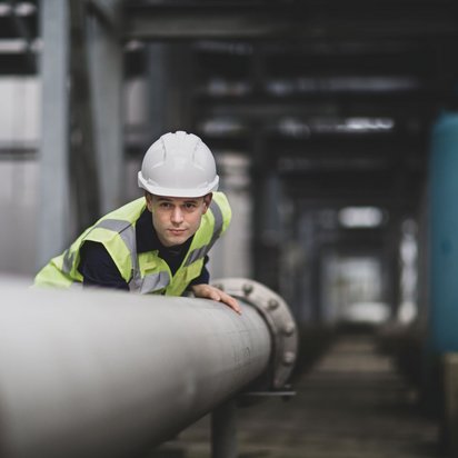 Person mit weißem Schutzhelm und gelber Warnweste, die sich in einer Industrieanlage befindet. Die Person lehnt sich über ein großes Metallrohr. 