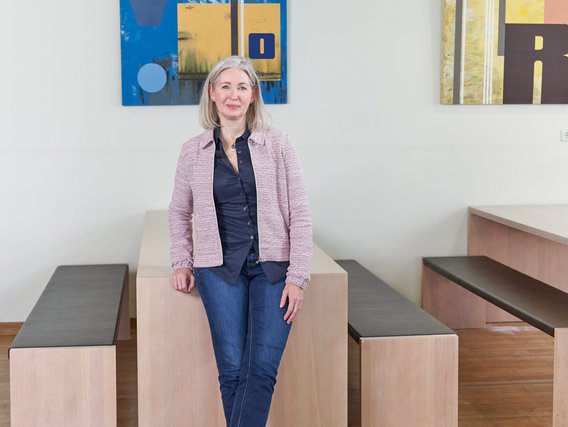 Mary Anderson Mary Anderson steht vor einem Tisch mit zwei Bänken. Hinter ihr an der Wand hängen Bilder mit abstrakter Kunst.