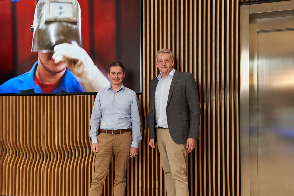 Carsten Gelhard und Knur Hirsch stehen vor einer Wand mit Holzelementen