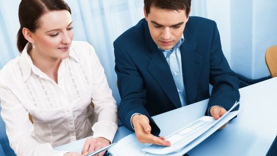 Versicherungen Eine Frau und ein Mann sitzen in Businesskleidung nebeneinander an einem Tisch. Sie schauen gemeinsam auf ein Blatt Papier. Vor der Frau liegt ein Ordner mit weiteren Dokumenten.