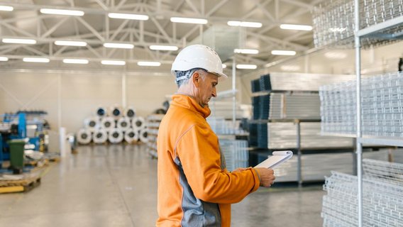 Mitarbeiter mit Schutzhelm kontrolliert Notizen in einer Produktions- oder Lagerhalle.