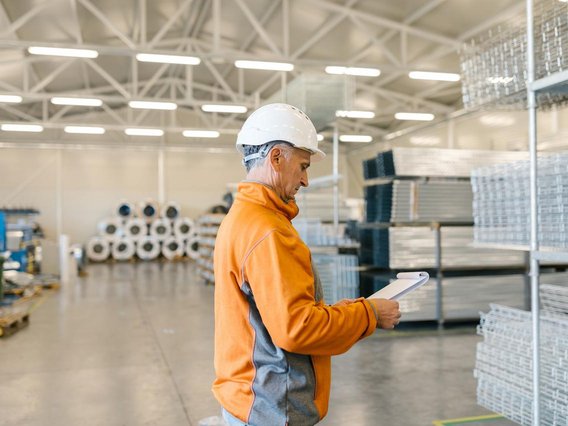 Arbeiter_Industriehalle Mitarbeiter mit Schutzhelm kontrolliert Notizen in einer Produktions- oder Lagerhalle.