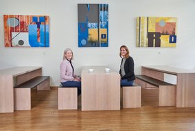 Mary Anderson und Natalie Morris sitzen an einem Tisch. Auch dem Tisch stehen zwei Tassen.