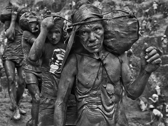 Mineradores Mineiros carregados com sacos caminham por uma trilha.