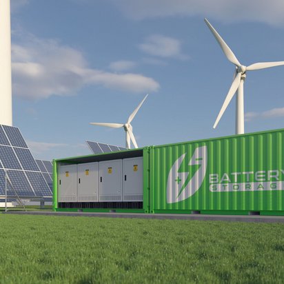 grünen Batteriespeicher in einem Container, der auf einer Wiese steht. Im Hintergrund sind mehrere große Solarmodule und Windkraftanlagen zu sehen. 