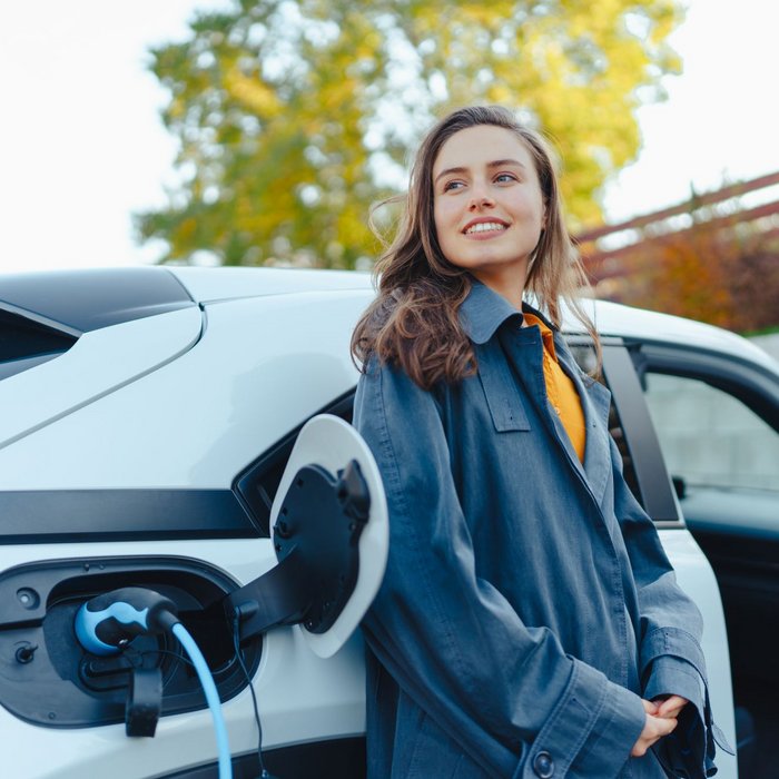 Eine junge Frau steht neben einem weißen Elektroauto, das gerade an einer Ladestation aufgeladen wird.