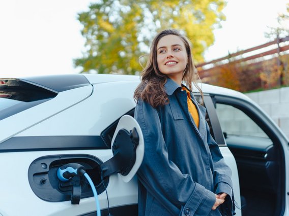 E_Mobilitaet_Person_Fahrzeug Eine junge Frau steht neben einem weißen Elektroauto, das gerade an einer Ladestation aufgeladen wird.