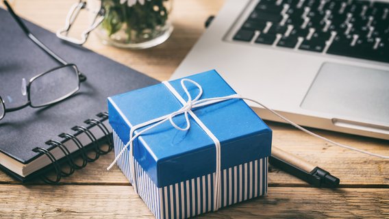 Geschenk Eine blaue Geschenkbox liegt auf einem Schreibtisch. Drumherum ist Notizblock, ein Laptop, ein Stift und eine Brille.
