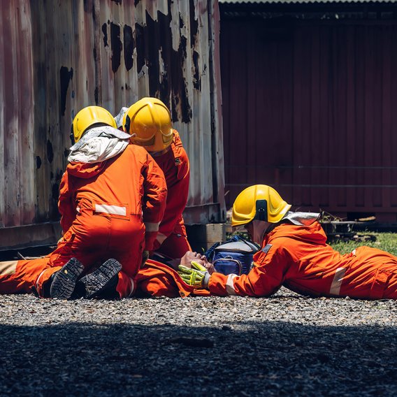 Drei Notfallsanitäter in orangenen Overalls bei einer Einsatzübung und dem Versuch, einen jungen Menschen auf einer Trage, die auf dem Boden steht, medizinisch zu versorgen.